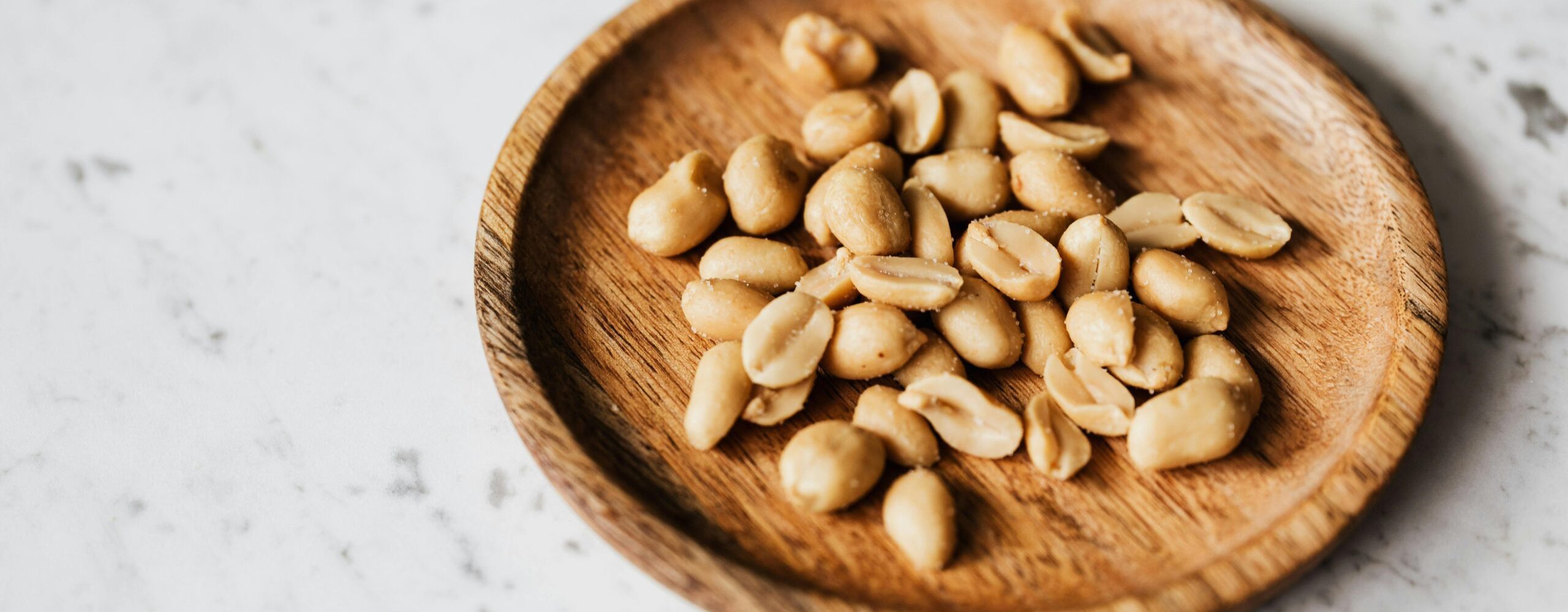 Top view of roasted peanuts in a wooden plate placed on a marble surface.