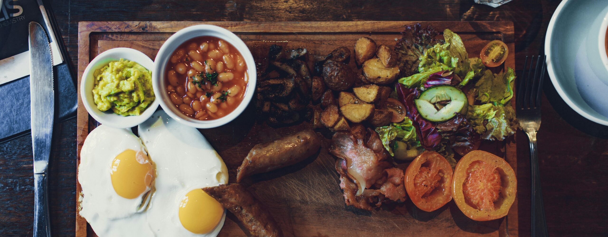 Top view of delicious fried eggs and vegetables with sausages and beans on wooden tray with fork and knife