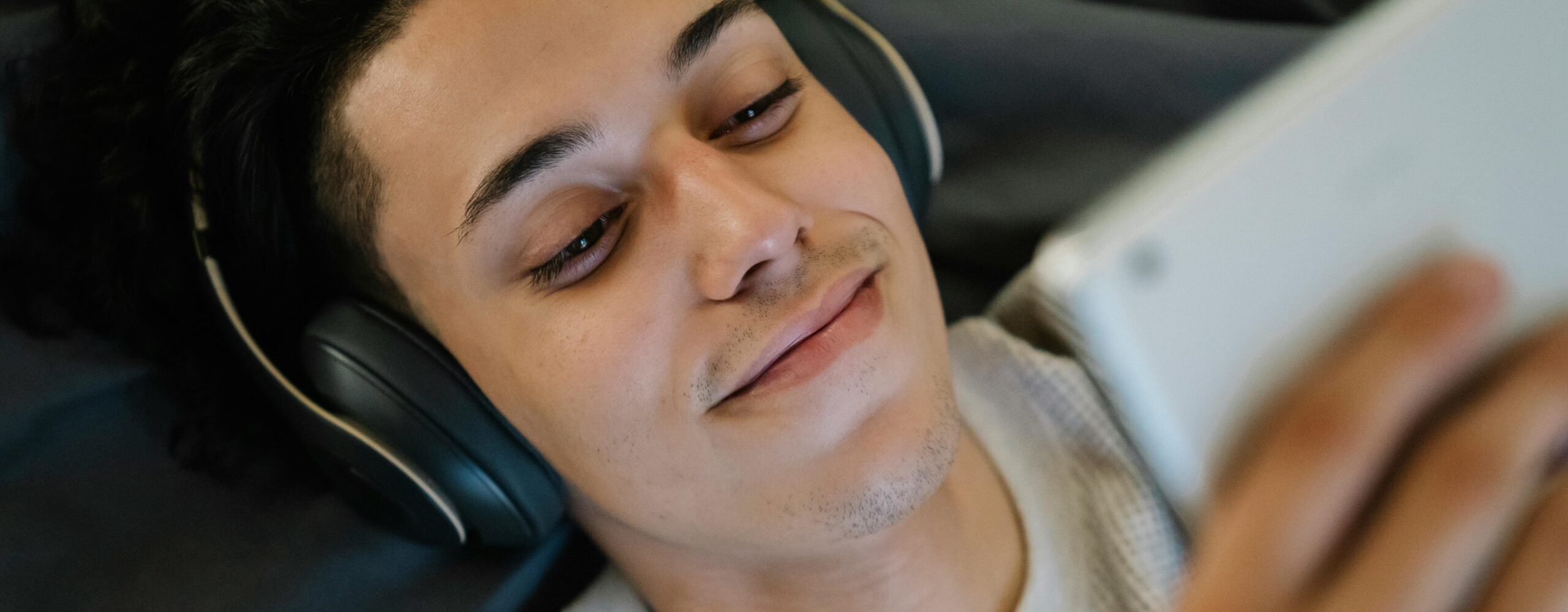 Positive young Hispanic man in headphones smiling and watching film on tablet while relaxing on bed at home