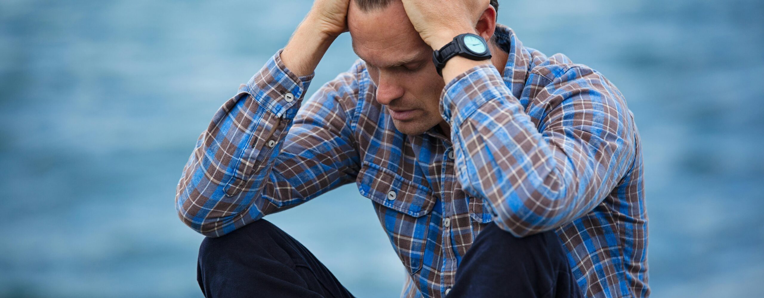 A man in a plaid shirt sits by the water looking distressed, symbolizing stress.